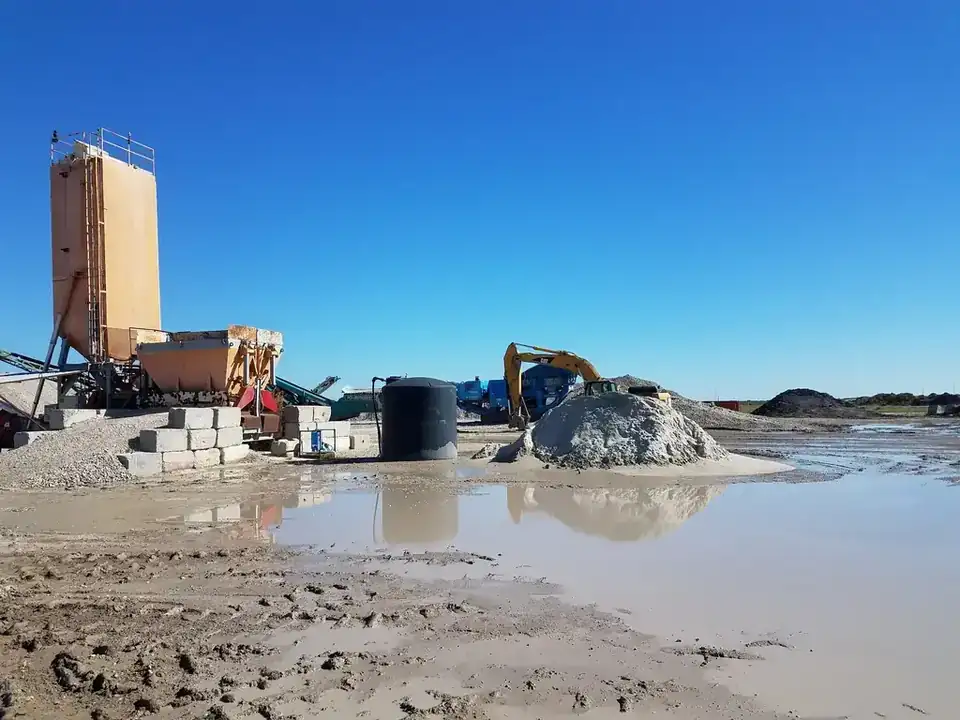 The Aaron Concrete Aransas Pass yard, a silo and Powerscreen crusher behind a CAT excavator and crushed stockpiles reflected in standing water