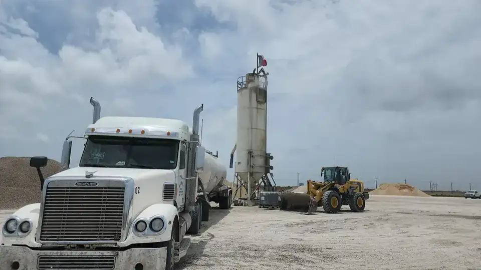 A Freightliner cement tanker beside a silo flying a Texas flag and a Komatsu loader