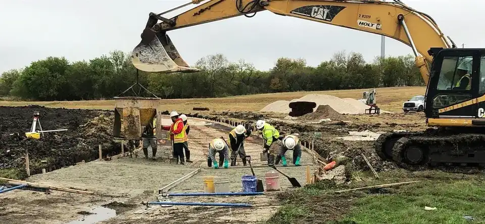 An Aaron Concrete crew hand-finishing a formed concrete slab with a survey tripod set and a CAT excavator working behind them