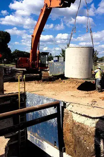 An excavator setting a precast manhole barrel over a trench box