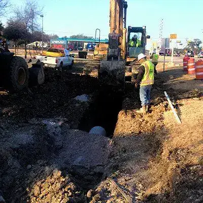 An excavator digging a utility trench in a street, pipe exposed and cones out